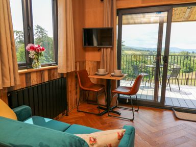 A small dining area with two chairs and a table set with bowls and plates next to a sliding glass door leading to a balcony overlooking green fields and hills at Rooster's Rest in Rhaedr Gwy near Rhayader