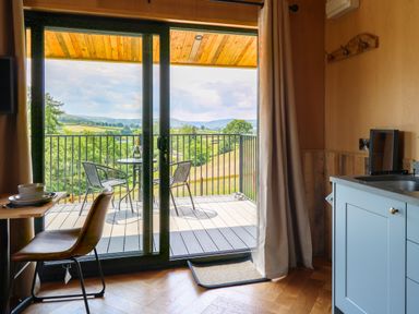 A kitchen area with blue cabinets and a sink next to a dining table with a chair by a sliding glass door opening to a balcony with two chairs and a table overlooking a landscape at Rooster's Rest in Rhaedr Gwy near Rhayader