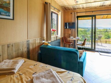 A room with a bed and towels a teal sofa a small dining table with two chairs a wall-mounted TV and a balcony overlooking a green landscape at Rooster's Rest in Rhaedr Gwy near Rhayader