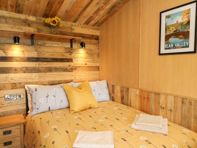 A bedroom with a wooden paneled wall a bed with floral bedding and pillows a wooden shelf with flowers and framed artwork at Rooster's Rest in Rhaedr Gwy near Rhayader