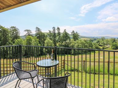 A balcony with two chairs and a table with a bottle and glasses overlooking trees and fields at Rooster's Rest in Rhaedr Gwy near Rhayader