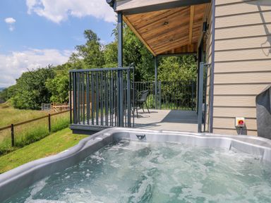An outdoor hot tub next to a covered deck with table and chairs overlooking green fields and trees at Rooster's Rest in Rhaedr Gwy near Rhayader