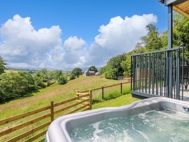 A hot tub on a deck overlooking a grassy field with a wooden fence and a house in the distance at Rooster's Rest in Rhaedr Gwy near Rhayader