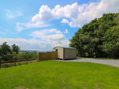 A small shed on a grassy hill with trees and a wooden fence under a cloudy blue sky at Rooster's Rest in Rhaedr Gwy near Rhayader