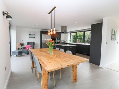 A dining area with a wooden table and grey chairs next to a kitchen with black cabinets and large windows at Pen-Y-Bryn in Llanfairpwllgwyngyll