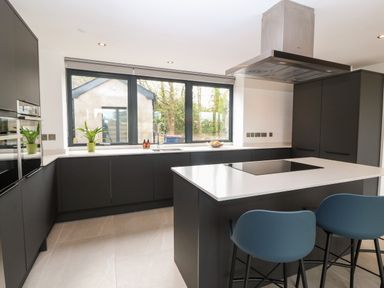 A kitchen with a large window black cabinets an island with a cooktop and two blue chairs at Pen-Y-Bryn in Llanfairpwllgwyngyll