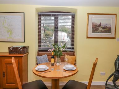 A dining area with a round wooden table set for two people near a window with blinds and framed artwork on walls at Bradley Stables in Withypool