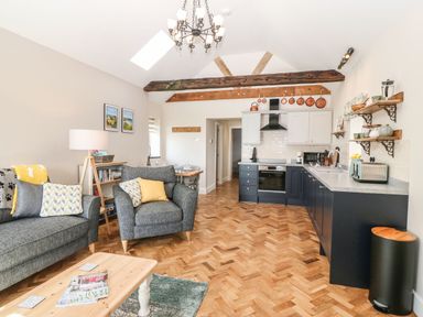 An open plan living area with grey armchairs and a sofa a wooden coffee table and a kitchen with black lower cabinets and cream upper cabinets at Pip's Barn in Brynteg near Benllech