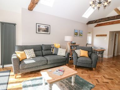 A living room with a gray sofa and armchair with yellow and patterned cushions a wooden coffee table a floor lamp and a dining table at Pips Barn in Brynteg near Benllech