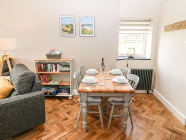 A dining area with a wooden table set for four and a small bookshelf near a gray sofa at Pips Barn in Brynteg near Benllech
