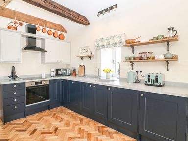 A kitchen with dark lower cabinets, light upper cabinets, wooden floor, shelves with cups and jars, and kitchen appliances at Pips Barn in Brynteg near Benllech