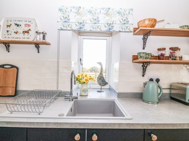 A kitchen sink with a drying rack cutting board kettle and toaster with shelves holding cups jars and a window with a bird ornament at Pips Barn Brynteg near Benllech