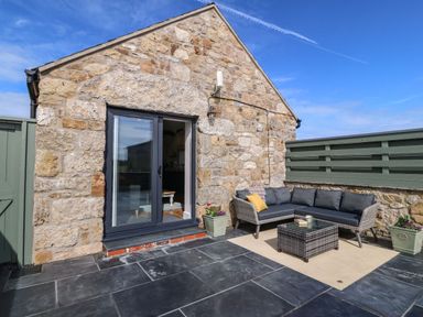 An outdoor patio area with a stone wall building glass door a grey sectional sofa with cushions a wicker table and flower pots at Pip's Barn in Brynteg near Benllech