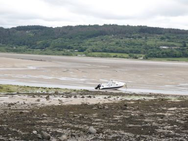 A small boat stranded on a sandy and rocky shore with hills and trees in the background at Pips Barn in Brynteg near Benllech