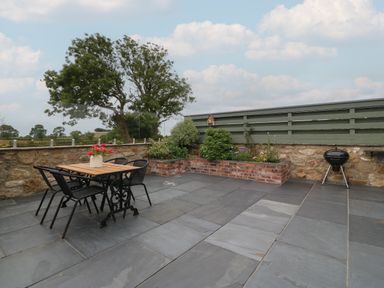 An outdoor patio with a wooden table and black chairs a small garden bed a barbecue grill and a green fence at Pip's Barn in Brynteg near Benllech