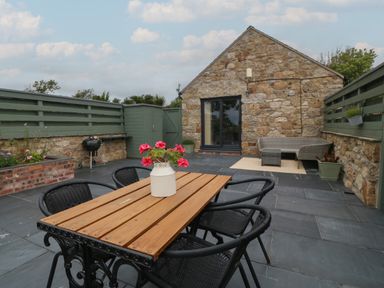 An outdoor patio area with a wooden table and black chairs a stone building and a small barbecue at Pips Barn in Brynteg near Benllech