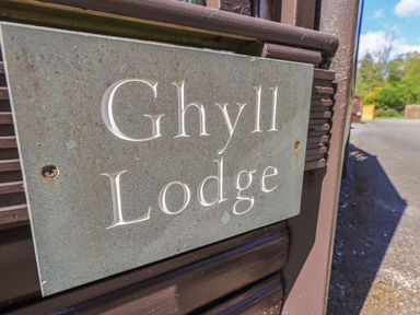 A close-up of a metal sign with the text Ghyll Lodge attached to a wooden post with a road and trees in the background at Ghyll Lodge Grasmere 25 White Cross Bay Windermere