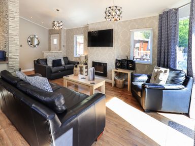 A living room with black leather sofas wooden tables a wall-mounted TV and windows at Ghyll Lodge Grasmere 25 White Cross Bay Windermere