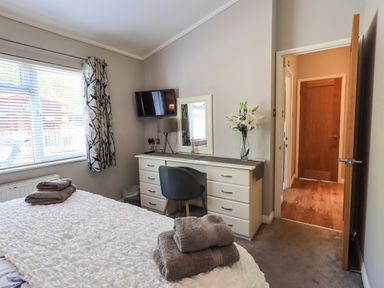 A bedroom with a bed and towels on it a desk with a chair a TV on the wall and a door leading to a hallway at Ghyll Lodge Grasmere 25 White Cross Bay Windermere