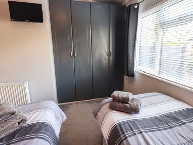 A bedroom with two single beds, a wall-mounted TV, a radiator, a corner wardrobe, and a window with blinds at Ghyll Lodge Grasmere 25 White Cross Bay Windermere
