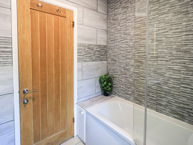 A bathroom with a wooden door a bathtub with glass panel and tiled walls with a small green plant at Ghyll Lodge Grasmere 25 White Cross Bay Windermere