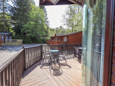 An outdoor wooden deck with metal chairs and tables surrounded by trees and wooden cabins at Ghyll Lodge Grasmere 25 White Cross Bay Windermere
