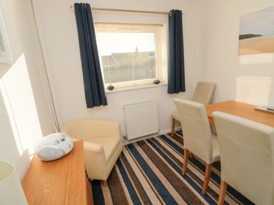 A dining room with a wooden table and cream chairs a cream armchair a wooden cabinet with a radio a window with dark curtains and blinds at Barnacles in Boulmer near Longhoughton