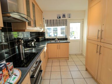 A kitchen with wooden cabinets a black tiled backsplash tiled floor a sink under a window and kitchen appliances at Barnacles in Boulmer near Longhoughton
