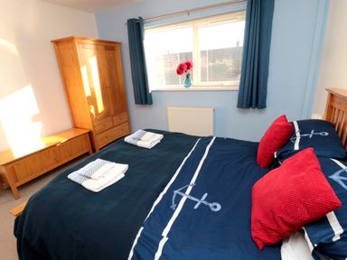 A bedroom with a bed with navy blue bedding with anchors and red pillows a wooden wardrobe a wooden bench under a window with blue curtains and a vase with red flowers at Barnacles Boulmer near Longhoughton