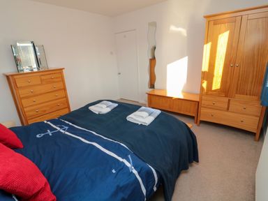 A bedroom with a bed covered in blue bedding red pillows wooden wardrobe chest of drawers and a wall mirror at Barnacles Boulmer near Longhoughton