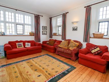 A seating area with four red sofas a patterned rug wooden floor and windows with curtains at Wick Court Farm in Arlingham near Frampton On Severn