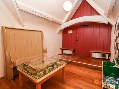A room with wooden floors a wooden bench a red wooden wall with two radiators a table with a display case containing a model at Wick Court Farm in Arlingham near Frampton On Severn