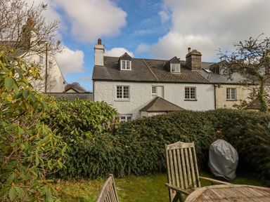 A garden with wooden chairs and a covered barbecue grill in front of a row of white and beige houses with slate roofs at Early Mist Cottage in Haytor Vale