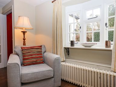 A living room corner with a gray armchair and a striped cushion a lamp and a bay window with a radiator and decorative items Early Mist Cottage Haytor Vale