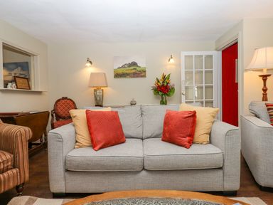 A living room with a gray sofa and red and beige cushions a striped armchair a wooden chair a lamp a flower vase a painting on the wall and a glass-paneled door at Early Mist Cottage in Haytor Vale
