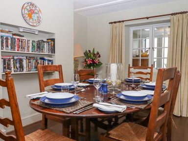 A dining room with a wooden table set for six with plates glasses and cutlery near a window and a bookshelf filled with DVDs at Early Mist Cottage in Haytor Vale