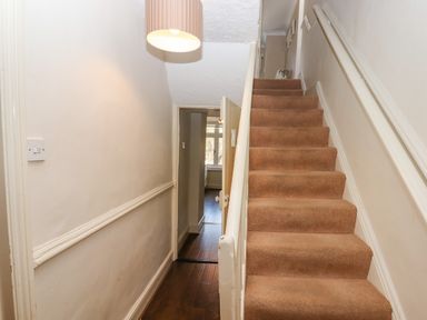 A hallway with a staircase covered in brown carpet and a doorway leading to a room with a window at Early Mist Cottage in Haytor Vale
