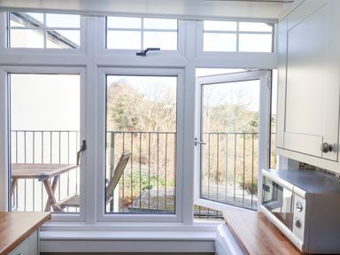 A kitchen window overlooking a balcony with a wooden table and chair and trees in the background at Early Mist Cottage in Haytor Vale