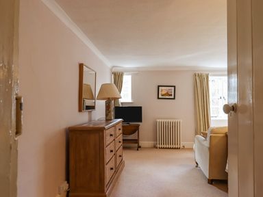 A living room with a wooden dresser with a lamp a wall mirror a small TV on a table a radiator two windows with curtains and a sofa at Early Mist Cottage in Haytor Vale