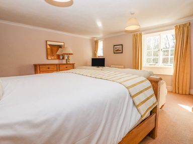 A bedroom with a wooden bed a dresser lamp mirror television and windows with curtains at Early Mist Cottage in Haytor Vale