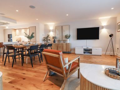 An open plan living and dining area with a wooden dining table and black chairs a leather armchair a wall mounted TV and wooden flooring at 7 Longshore in St Columb Minor