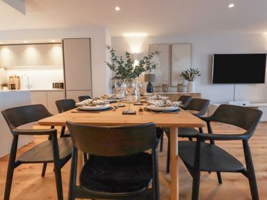 A dining table set with plates glasses and napkins in a room with chairs kitchen and wall TV at 7 Longshore in St Columb Minor