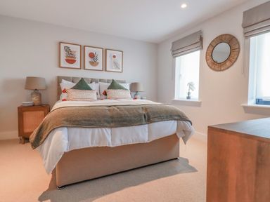 A bedroom with a bed with pillows and a blanket bedside tables with lamps wall art a circular mirror and windows at 7 Longshore in St Columb Minor
