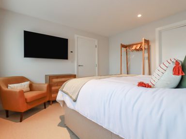 A bedroom with a bed, a mounted television, a wooden chest, an armchair, and a wooden clothes rack at 7 Longshore in St Columb Minor