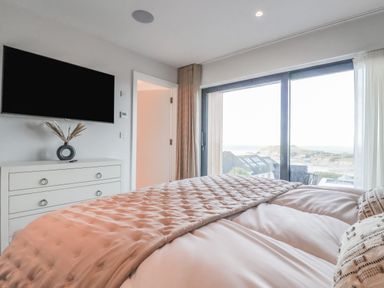 A bedroom with a bed facing a TV mounted on the wall and a large window showing an outdoor view at 7 Longshore in St Columb Minor