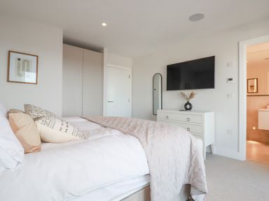 A bedroom with a bed pillows a blanket a chest of drawers a wall-mounted tv a mirror a wardrobe and a door leading to a bathroom at 7 Longshore in St Columb Minor