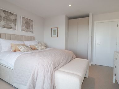 A bedroom with a large bed pillows and cushions a bench at the foot of the bed a wardrobe a chest of drawers and abstract artwork on the walls at 7 Longshore in St Columb Minor
