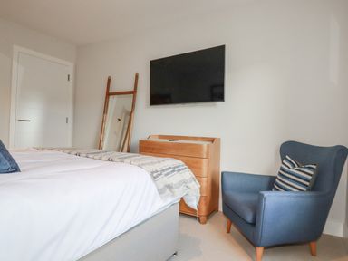 A bedroom with a bed a wooden dresser a wall mounted tv a floor mirror and a blue armchair at 7 Longshore in St Columb Minor