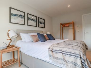 A bedroom with a bed pillows a blanket bedside table a lamp and a wooden clothes rack at 7 Longshore in St Columb Minor
