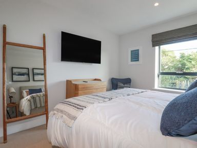 A bedroom with a bed a wooden dresser a mounted TV a window and a wooden floor mirror at 7 Longshore in St Columb Minor